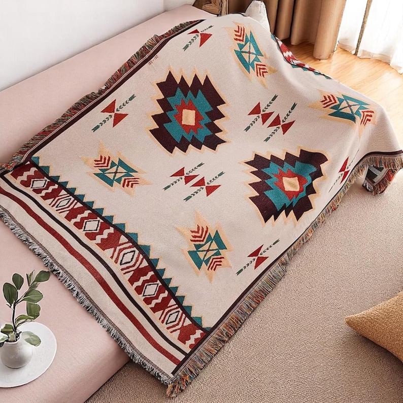 May include: Beige woven throw blanket with a geometric pattern in teal, burgundy, and cream. The blanket has a fringed edge and is draped over a pink sofa. The design features repeating shapes and patterns.