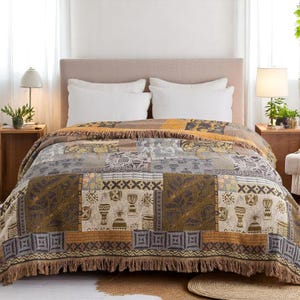 Puede incluir: Una manta de estilo patchwork con borde de flecos, que presenta una mezcla de patrones geométricos y tribales en tonos marrón, beige, gris y dorado. La manta está colocada sobre una cama con almohadas blancas y un cabecero beige.