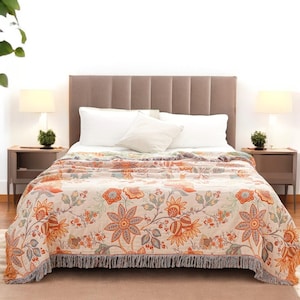 Op de afbeelding: Een beige bloemen deken met oranje en blauwe accenten, gefranjerde randen en een herhalend bloemmotief. De deken is gedrapeerd over een bed met witte kussens en een bruin hoofdbord. Twee lampen staan aan weerszijden.
