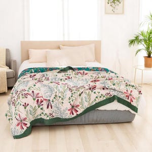 Op de afbeelding: Een bloemenpatroon deken met een crèmekleurige achtergrond en een groene rand is over een bed gedrapeerd. De deken heeft een ontwerp van roze en paarse bloemen, groene bladeren en blauwgrijze stelen. Het bed heeft witte lakens en kussens.