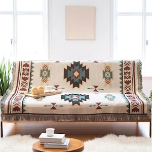 May include: A beige sofa with a patterned throw blanket. The blanket features geometric designs in shades of brown, teal, and red. The sofa has a wooden tray with bread and a cup. The fringe on the blanket adds a decorative touch.