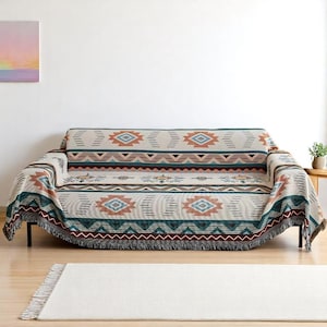 May include: A beige sofa throw blanket with a geometric pattern in shades of teal, rust, and brown. The throw has a fringed edge and is draped over a black sofa. A white rug is on the floor.