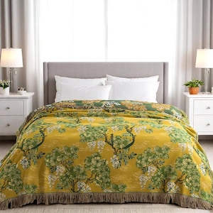 May include: A yellow and green throw blanket with a botanical design featuring trees and flowers. The blanket has a fringed edge and is draped over a bed. The colour palette includes shades of yellow, green, and white.