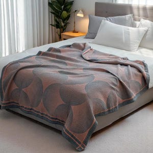 Pode incluir: Uma colcha com um padrão geométrico de círculos cinza e pêssego sobrepostos. O cobertor tem uma borda cinza com uma faixa pêssego. O cobertor é colocado sobre uma cama com travesseiros brancos.