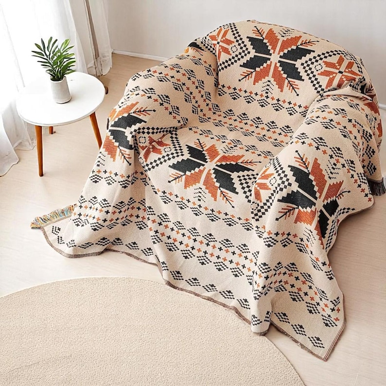 May include: A beige throw blanket with a geometric pattern of black, orange, and brown shapes draped over a chair. The blanket has a fringed edge and is made of a woven material. A small side table with a plant is visible in the background.