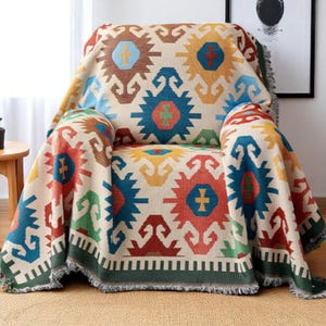 May include: A beige throw blanket with a colourful geometric pattern covers an armchair. The pattern includes shapes in red, blue, yellow, and brown. The blanket has a green border and fringe.