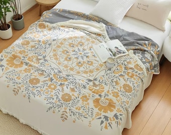 May include: A floral patterned blanket in cream, gray, and gold covers a bed. The design features large stylized flowers and leaves. The blanket has a decorative border and is paired with white pillows. A book and candle sit on top.