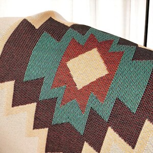 May include: Close-up of a woven blanket with a geometric pattern. The design features a central diamond shape in beige, surrounded by red, teal, and dark brown zigzags and diamond shapes. The blanket has a beige background.