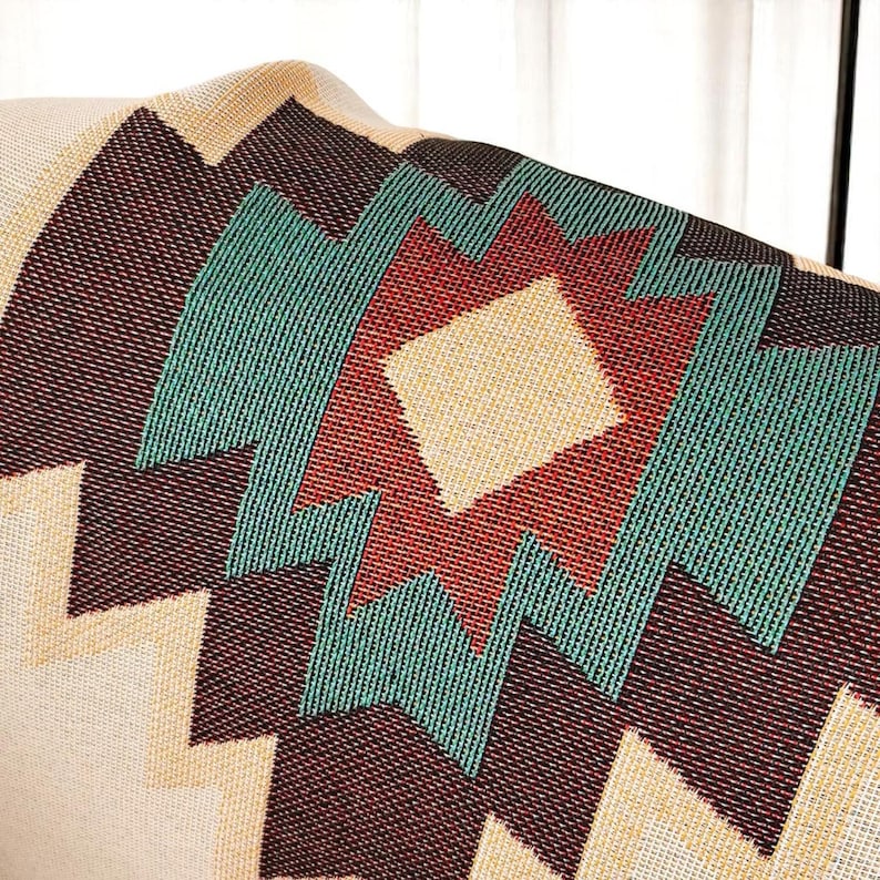 May include: Close-up of a woven blanket with a geometric pattern. The design features a central diamond shape in beige, surrounded by red, teal, and dark brown zigzags and diamond shapes. The blanket has a beige background.