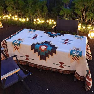 May include: A rectangular cream-colored tablecloth with a geometric pattern in teal, burgundy, and brown. The tablecloth has a fringed edge and is draped over a table. Two folding chairs are visible.