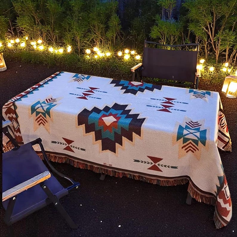 May include: A rectangular cream-colored tablecloth with a geometric pattern in teal, burgundy, and brown. The tablecloth has a fringed edge and is draped over a table. Two folding chairs are visible.