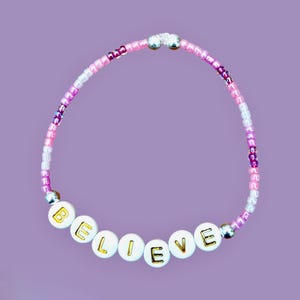 Könnte beinhalten: Ein Perlenarmband mit dem Wort "BELIEVE" in goldenen Buchstaben auf weißen Perlen. Das Armband hat rosa, lila und klare Perlen mit silbernen Akzenten. Der Hintergrund ist einfarbig lavendelfarben.