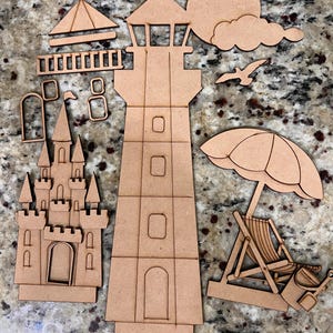 May include: A collection of unfinished light brown wooden cutouts featuring a tall lighthouse, a detailed castle, a beach umbrella with a lounge chair, a sun, a cloud, a seagull, and various small architectural components for assembly.