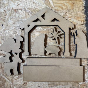 May include: A set of unfinished light brown wooden nativity scene cutouts including a stable structure, a star, figures of people, a camel, and a donkey, accompanied by a rectangular wooden base piece for assembly and display purposes.