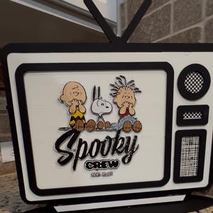 May include: A black and white decorative TV featuring the Peanuts characters Charlie Brown, Snoopy, and Pig-Pen. The words "Spooky Crew" are written across the front of the TV, with the phrase "Good Grief!" below.