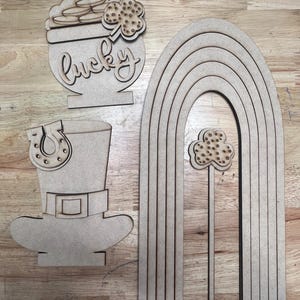 May include: Wooden cutouts for St. Patrick's Day decor. Includes a pot of gold with the word "lucky", a leprechaun hat with a horseshoe, and a rainbow with a shamrock. All pieces are made of unfinished wood.