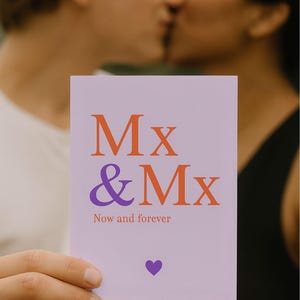 Könnte beinhalten: Eine lila Grußkarte mit dem Text "Mx & Mx" in Orange und Lila, darunter "Now and forever" und ein lila Herz. Die Karte wird vor zwei sich küssenden Personen gehalten.