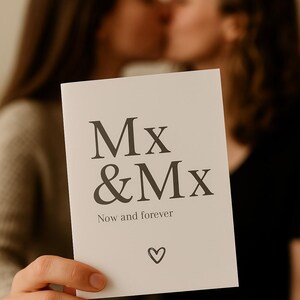 Könnte beinhalten: Eine weiße Karte mit dem Text "Mx & Mx" und "Now and forever" in einer Serifenschrift, mit einem Herzsymbol. Die Karte wird vor zwei sich küssenden Personen gehalten, die im Hintergrund unscharf sind.