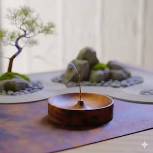 以下が含まれることがあります： 木製の香立てに、煙を出す燃えているお香が置かれています。ホルダーは茶色の表面に置かれ、背景には岩、砂、小さな盆栽を備えたミニチュアの禅ガーデンがあります。このシーンは静けさを呼び起こします。