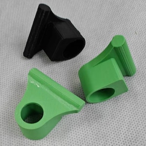 Könnte beinhalten: Drei 3D-gedruckte Kunststoffobjekte. Eines ist schwarz, zwei sind grün. Jedes hat eine ringförmige Basis und eine abgewinkelte, rechteckige Oberseite. Die Objekte scheinen klein zu sein, möglicherweise Komponenten oder Zubehör.