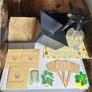 Pode incluir: Um kit de cultivo de plantas com uma jardineira cinza escuro, um borrifador, saquinhos de sementes e marcadores de plantas. O kit inclui adesivos de plantas e um cartão com o texto "Growing With Us!" e "Botanikit".