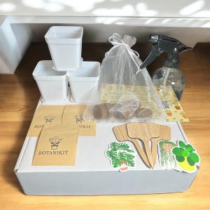 May include: A white box containing a Botanikit, with white pots, a spray bottle, seed packets, and plant markers. A sheer bag holds brown seed starters. Decorative plant stickers are also included.