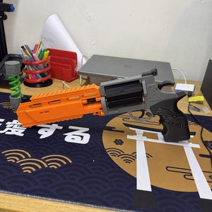 Könnte beinhalten: Ein Spielzeugrevolver mit einem leuchtend orangefarbenen Lauf und einer oberen Schiene, einer schwarzen Trommel, einem grauen Rahmen und einem schwarzen Griff mit texturiertem Flügeldesign. Das Spielzeug besteht aus verschiedenfarbigen Kunststoffteilen.