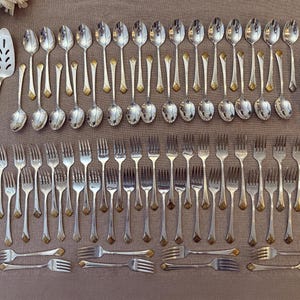 Pode incluir: Uma coleção de talheres prateados, incluindo colheres, garfos e utensílios de servir, dispostos sobre uma superfície de tom neutro. A prataria tem detalhes dourados nas alças. Flores brancas ao fundo.