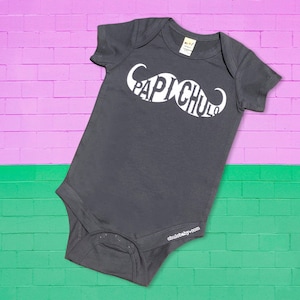 May include: A dark gray baby onesie with a white mustache design that says "Papichulo" on it. The onesie has short sleeves and snaps at the crotch. The onesie is on a pink and green brick background.