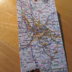 Op de afbeelding: Een rechthoekig label gemaakt van een kaart van Pittsburgh, Pennsylvania, met een bruin lint bevestigd. Het label heeft een Pennsylvania-stempel en de woorden "HIT THE ROAD".