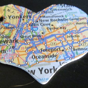 May include: Heart-shaped ornament featuring a map of the New York City area. The map includes cities like Newark, Yonkers, and Freeport. A tan ribbon is attached for hanging. The ornament is made of a map material.