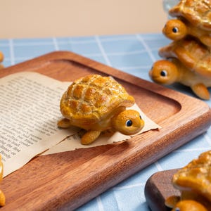 May include: A collection of decorative turtle figurines in shades of brown and yellow. One turtle sits on a piece of paper, while others are stacked or placed on a wooden tray. The turtles have detailed shell patterns and blue eyes.