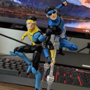Puede incluir: Dos figuras de acción de personajes de cómic, una con un traje azul y amarillo, la otra con un traje azul y negro. Ambas figuras están en poses dinámicas, con articulaciones y gafas de sol. Las figuras están sobre soportes transparentes, con un teclado y un monitor al fondo.