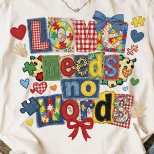 May include: White t-shirt with the phrase "LOVE needs no WORDS" in colorful, patchwork-style letters and shapes. The design includes hearts, puzzle pieces, and a bow. The shirt has a crew neck and short sleeves.