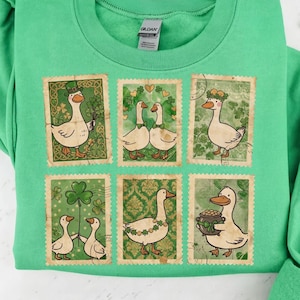 May include: Green sweatshirt with a design of six postage stamp-style images of cartoon geese with St. Patrick's Day themes. The geese wear hats, shamrocks, and hold pots of gold. The sweatshirt is a solid green colour.