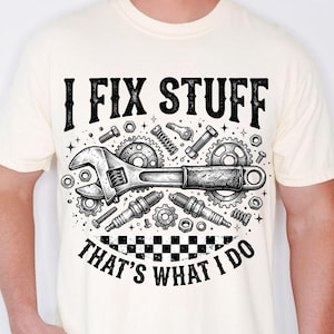 Puede incluir: Camiseta color crema con un gráfico en blanco y negro de herramientas, incluyendo una llave inglesa, engranajes y bujías. El texto "I FIX STUFF" está encima de las herramientas y "THAT'S WHAT I DO" debajo. El diseño está centrado.
