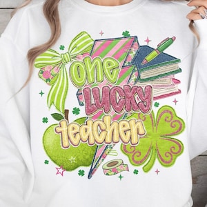 May include: White sweatshirt with a colorful graphic design. The design includes the text "One Lucky Teacher" in green and pink, along with a green apple, books, a shamrock, and a bow.