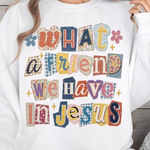 以下が含まれることがあります： カラフルなパッチワークスタイルの文字で「WHAT a FRIEND we HAVE in JESUS」と書かれた白いスウェットシャツ。文字は、花柄、ギンガムチェック、無地など、さまざまなパターンと色で表現されています。デザインには、小さな花と星のアクセントが含まれています。