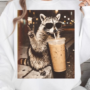 May include: White sweatshirt featuring a raccoon wearing sunglasses and holding an iced coffee. The raccoon is giving a peace sign. The image has a sepia tone, with a blurred background of city lights.