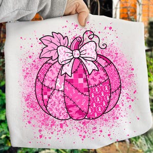 May include: A pink pumpkin design on a white background. The pumpkin is composed of various pink patterns, including ribbons, checks, and leopard print. A large bow and pink splatter complete the design. The image is held by a person.