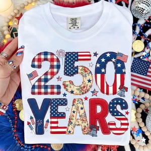 May include: White t-shirt with a patriotic design celebrating 250 years. The design features the number "250" and the word "YEARS" in red, white, and blue patterns, including the American flag and floral accents. The shirt has a crew neck.