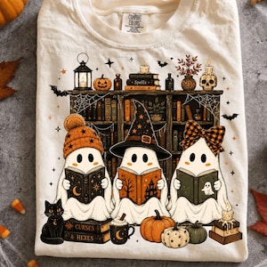 May include: A cream-colored t-shirt featuring a graphic of three ghosts reading books in front of a bookshelf. The ghosts wear an orange beanie, a black witch hat, and a plaid bow. The design includes pumpkins, a black cat, and candles.