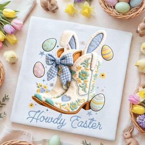 May include: White t-shirt with an embroidered design featuring cowboy boots with bunny ears, Easter eggs, and floral accents. The text "Howdy Easter" is embroidered in blue. The design incorporates light blue, yellow, and brown colors.