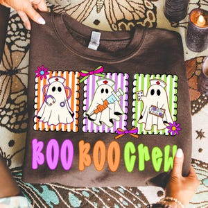 May include: A brown sweatshirt featuring three cartoon ghosts dressed as nurses, each in a different striped frame. The ghosts hold medical tools. The text "BOO CREW" is printed below in colorful letters. The sweatshirt is held by hands.