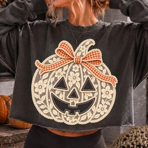 May include: A charcoal gray cropped sweatshirt featuring a large graphic of a pumpkin made from cream-colored lace with floral patterns. The pumpkin has black triangular eyes and a black mouth, topped with an orange and white gingham bow.
