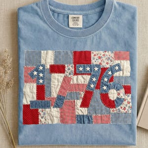 May include: Pale blue t-shirt with a patchwork design displaying the number "1776" in red, white, and blue. The design includes stars and patterns such as gingham and floral prints. The shirt has a crew neck and a small tag on the upper center.
