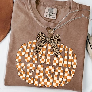May include: A brown short-sleeve t-shirt with a Comfort Colors label at the neck. The graphic features a pumpkin shape filled with an orange and white checkerboard pattern, topped with a leopard print bow and the text PUMPKIN SEASON.