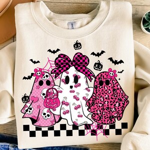 May include: Cream-colored sweatshirt featuring three ghost characters with pink accents. The ghosts have various patterns, including skulls, cherries, and leopard print. A large checkered bow and Halloween-themed decorations complete the design.