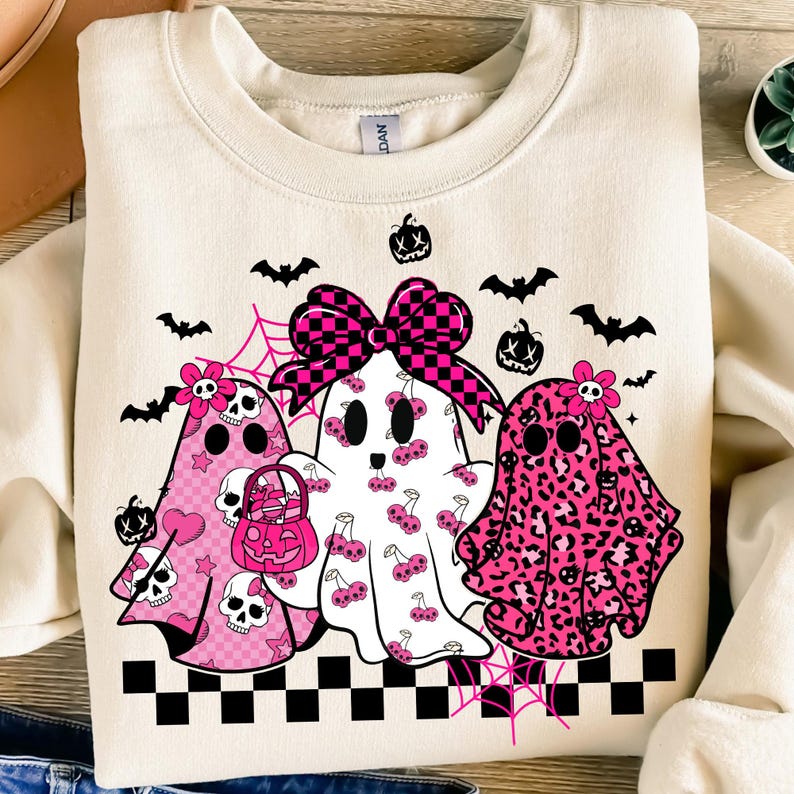 May include: Cream-colored sweatshirt featuring three ghost characters with pink accents. The ghosts have various patterns, including skulls, cherries, and leopard print. A large checkered bow and Halloween-themed decorations complete the design.