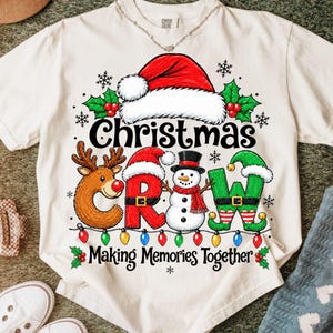 Op de afbeelding: Een crèmekleurig T-shirt met korte mouwen en een opdruk met de tekst "Christmas Crew" en "Making Memories Together". Het ontwerp bevat een rendier, een sneeuwpop, een kerstmuts, hulst en een snoer met kleurrijke kerstverlichting.